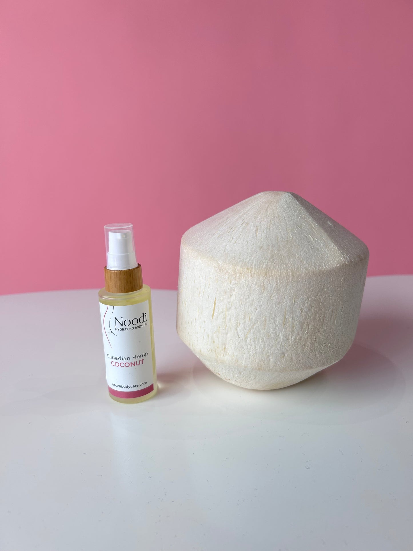 A bottle of Noodi Canadian Hemp Coconut body oil next to a decorative coconut shell, set against a pink background.