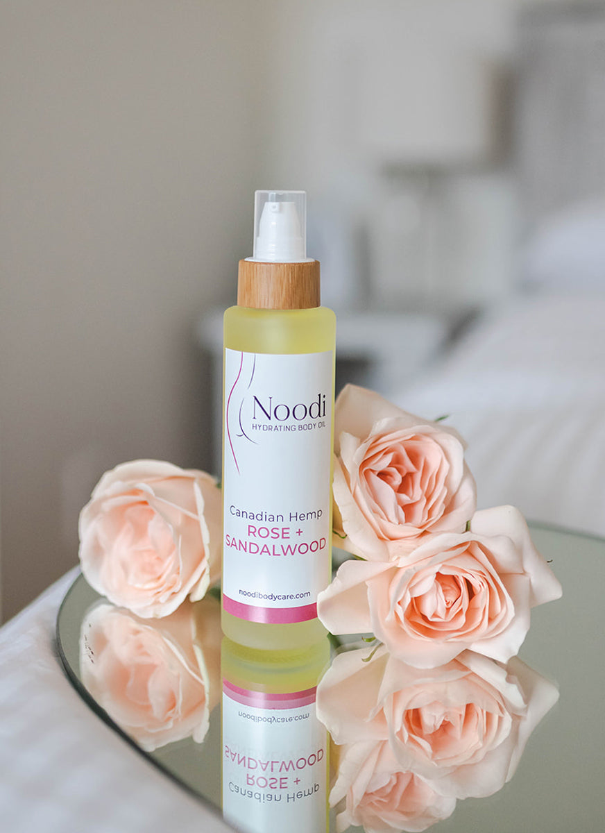 A bottle of Noodii Hydrating Body Oil, featuring Canadian Hemp, Rose, and Sandalwood, beside soft pink roses on a reflective surface.