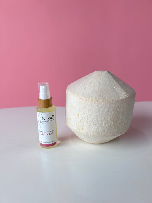 A bottle of Noodi Canadian Hemp Coconut body oil next to a decorative coconut shell, set against a pink background.