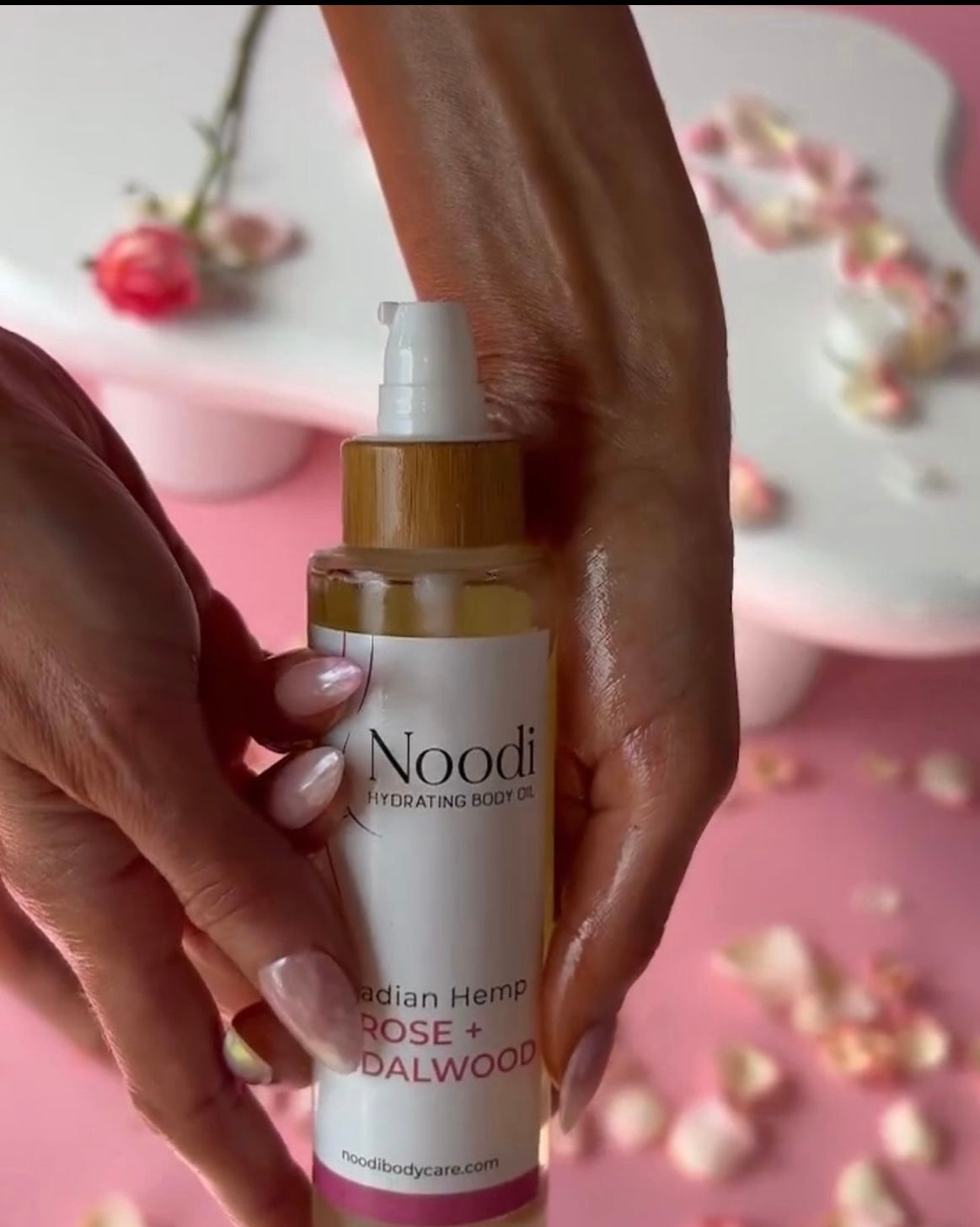 A hand holds a bottle of Noodi Hydrating Body Oil with a rose and sandalwood label, set against a pink background with flower petals.
