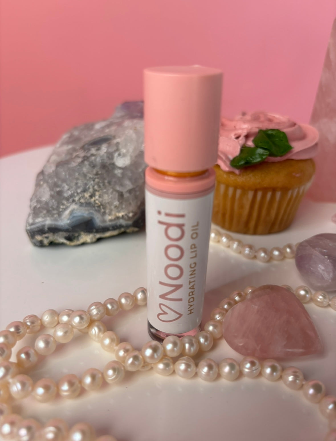 A pink-lidded lip oil tube is surrounded by pearls, crystals, and a frosted cupcake, set against a pink background.