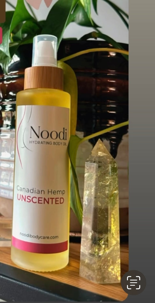 A bottle of Noodi Hydrating Body Oil next to a crystal, with a label reading "Canadian Hemp UNSCENTED," set against lush green foliage.