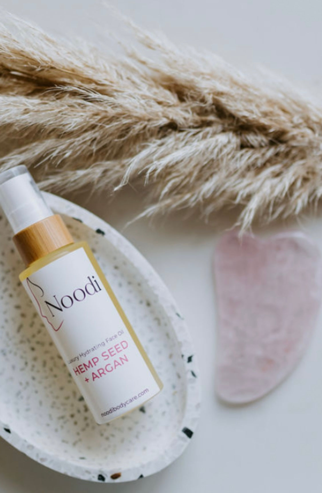 A bottle of Noodi hemp seed and argan face oil rests on a speckled dish with a rose quartz gua sha tool and dried pampas grass nearby.